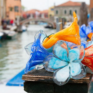 Vibrant Murano glass flowers in blue, orange, and red displayed on a wooden post beside a canal in Venice, with boats and colorful buildings in the background.