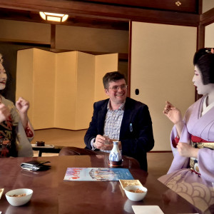 A man sits at a low table, smiling and conversing with two geisha dressed in traditional kimonos—one in blue and the other in lavender—inside a tatami room during a cultural experience, with drinks and snacks on the table.