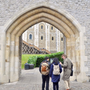 Tour group entering the Tower of London through a large stone archway, led by a guide in a trench coat.