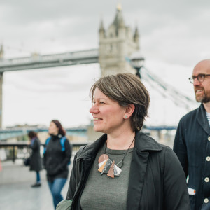 Two smiling individuals walking near Tower Bridge on a cloudy day, with the historic bridge in the background.