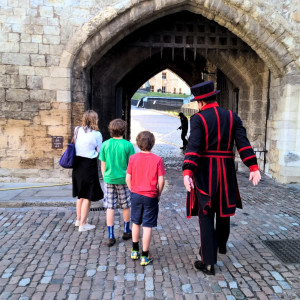 Beefeater leading a family through a stone archway inside the Tower of London, with cobblestone pathways and historic walls.