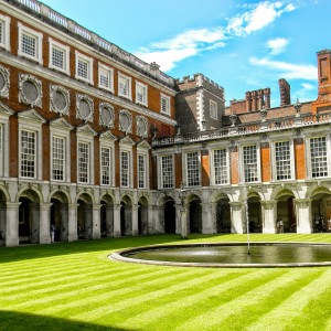 A courtyard at Hampton Court Palace with striped green lawn, circular fountain in the center, and red-brick walls with large arched windows surrounding the space.