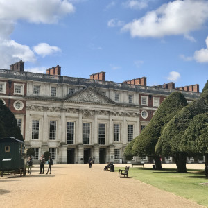 Front façade of Hampton Court Palace with tall windows, red brick and stone details, and visitors walking along the gravel path lined with shaped trees.