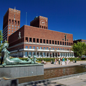 Red brick Oslo City Hall with statues and fountains in the foreground under a clear blue sky.