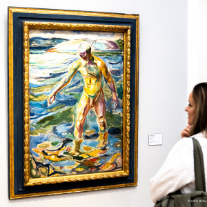 A woman observes a colorful framed painting of a figure standing in water.