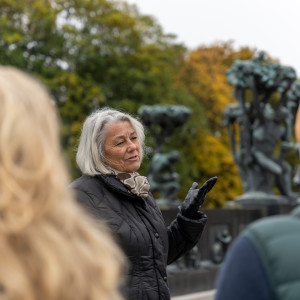 A woman speaks to visitors in front of bronze sculptures surrounded by autumn trees.