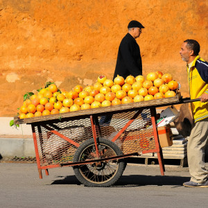 A street vendor in a yellow jacket pushes a wooden cart filled with oranges and pomegranates along a sunlit street in Morocco, with a man walking past a warm, terracotta-colored wall in the background.