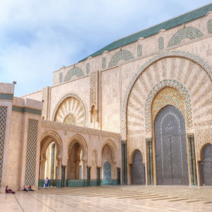 The ornate exterior of the Hassan II Mosque in Casablanca, featuring grand arched doorways, intricate geometric tilework, and towering carved stone facades under a pale blue sky.