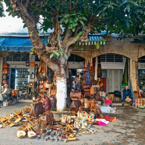 A row of artisan market stalls in Morocco shaded by large trees, displaying leather bags, rugs, metalwork, and handcrafted goods, with vendors seated among colorful items spread across the cobblestone ground.