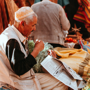 An older man wearing a patterned cap sits in a Marrakesh market stall, holding fresh herbs in one hand and reading a newspaper amid colorful sacks and produce.