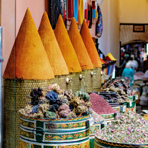 Tall, cone-shaped piles of spices line the front of a Marrakesh souk shop, surrounded by baskets of dried flowers, pigments, and herbs, with shoppers walking through the narrow passageway.