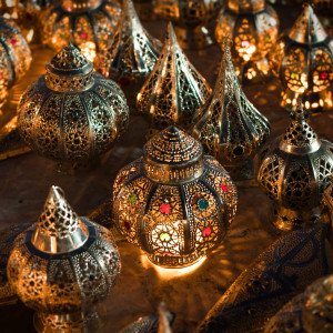 A display of intricately carved Moroccan metal lanterns glowing from within, their warm light illuminating colorful glass inlays and ornate geometric patterns.