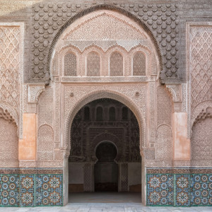 The ornate carved stone and zellij tilework of the Ben Youssef Madrasa in Marrakesh, featuring arched doorways, geometric patterns, and detailed stucco decoration.