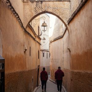 A narrow passageway in the Fez medina with high, sand-colored walls and arched stone supports, as two people walk through the winding lane beneath hanging lanterns.
