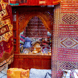 A vibrant Fes market stall displaying richly patterned Berber carpets and textiles, with carved wooden shelves filled with pottery, metalwork, and traditional crafts.