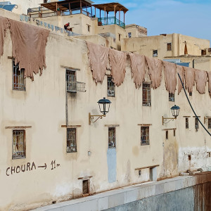 Large pieces of leather hang drying on the exterior walls of a building near the Chouara Tannery in Fez, with traditional lanterns and weathered windows beneath.
