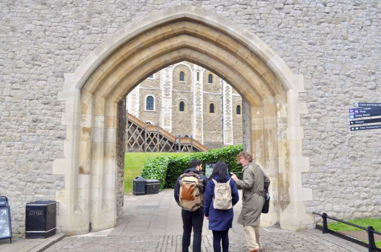 Tour group entering the Tower of London through a large stone archway, led by a guide in a trench coat.