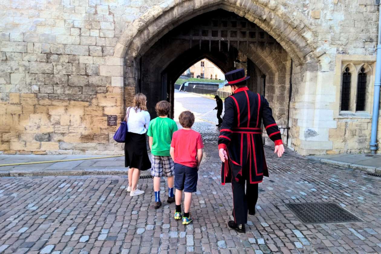 Beefeater leading a family through a stone archway inside the Tower of London, with cobblestone pathways and historic walls.