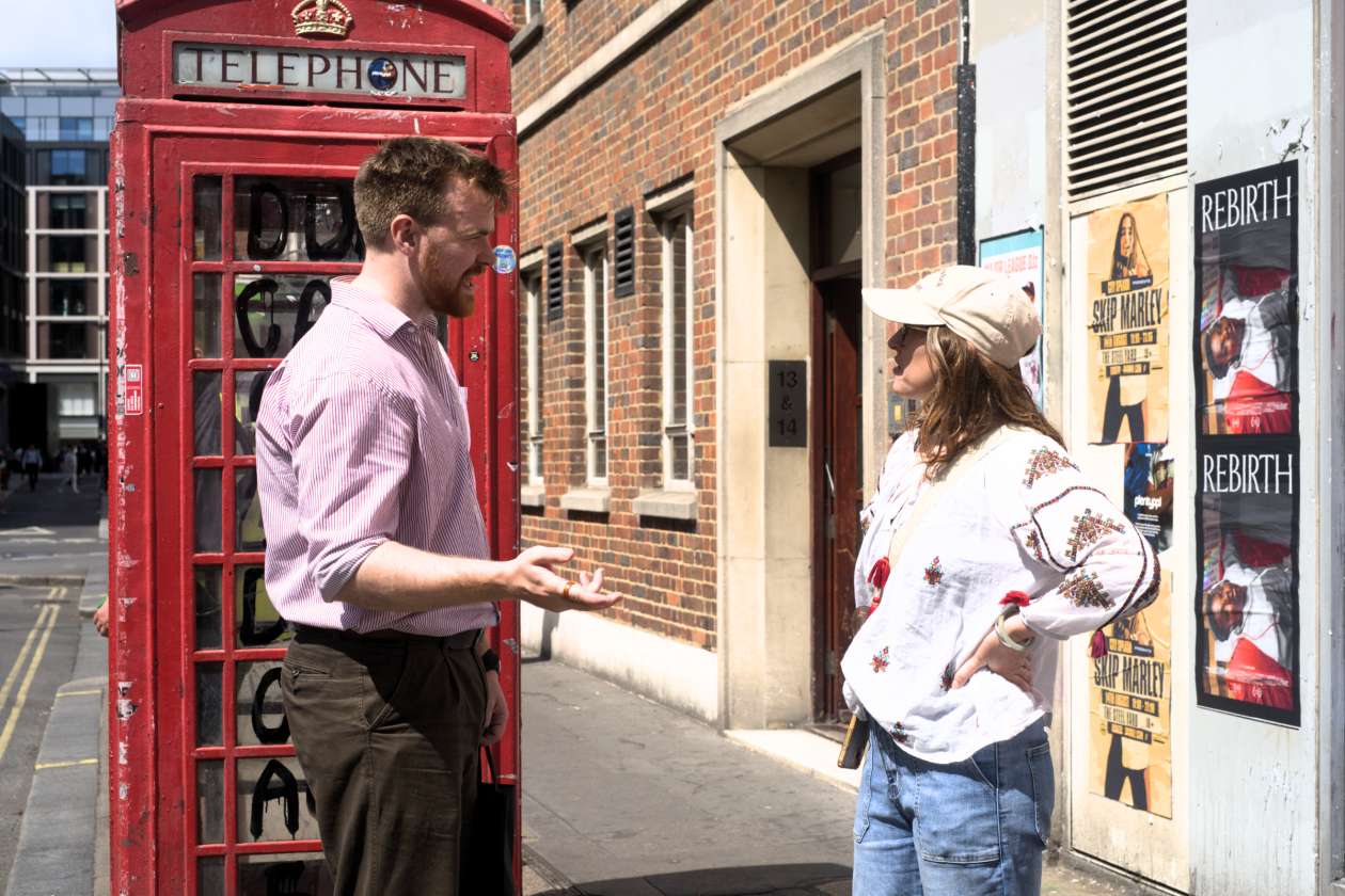 A guide speaks with a visitor beside a red telephone booth covered in graffiti in central London.