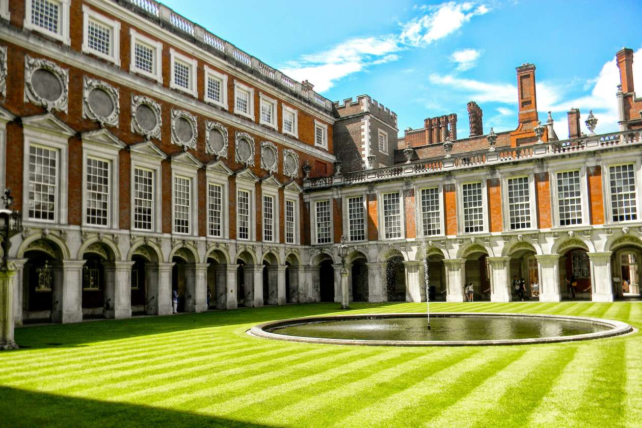 A courtyard at Hampton Court Palace with striped green lawn, circular fountain in the center, and red-brick walls with large arched windows surrounding the space.