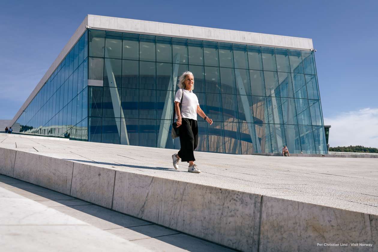 A woman walking along the sloped white marble roof of the Oslo Opera House on a sunny day.
