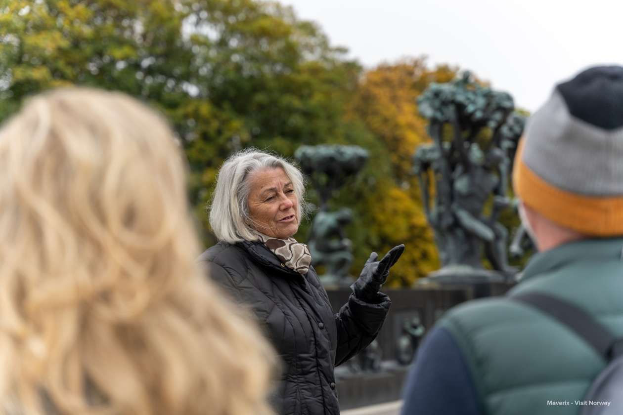 A woman speaks to visitors in front of bronze sculptures surrounded by autumn trees.
