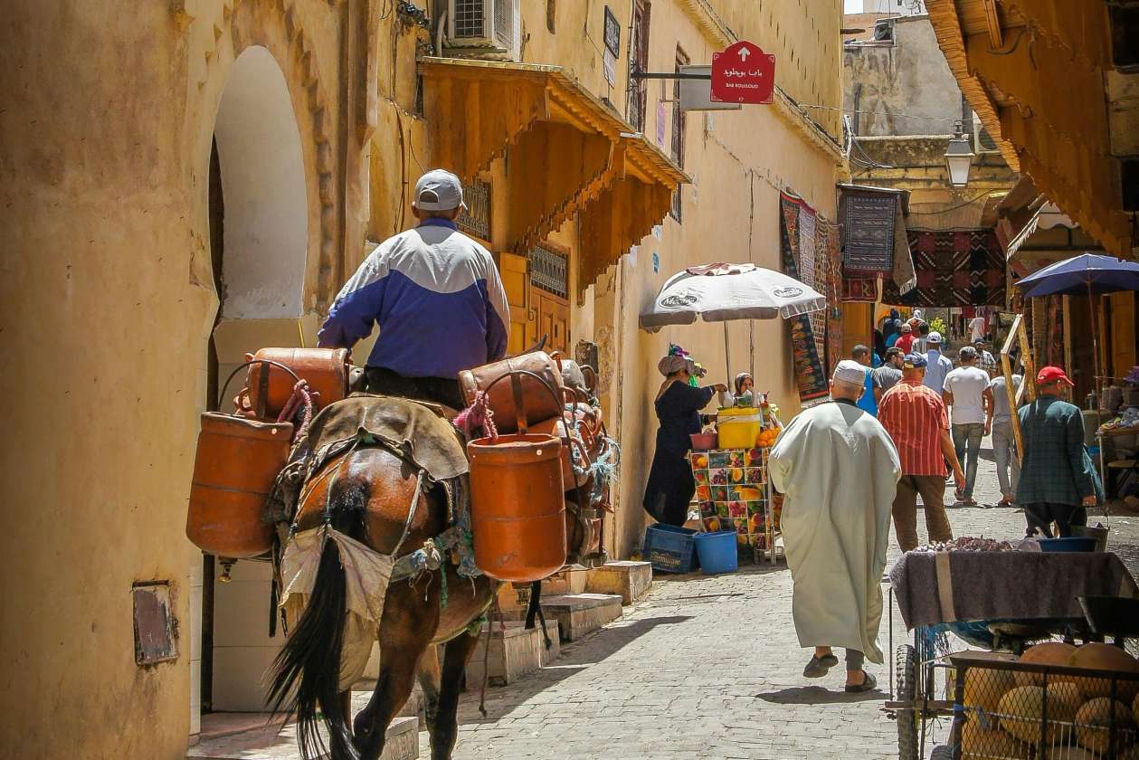 A busy medina alleyway where a man on a donkey carrying metal containers moves through a narrow street lined with shops, vibrant textiles, and locals walking under the warm, sand-colored buildings.