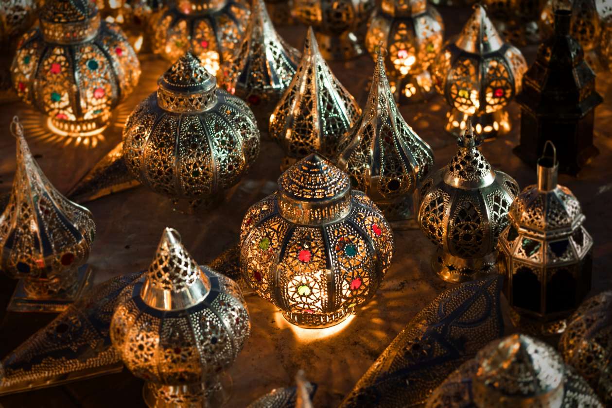 A display of intricately carved Moroccan metal lanterns glowing from within, their warm light illuminating colorful glass inlays and ornate geometric patterns.