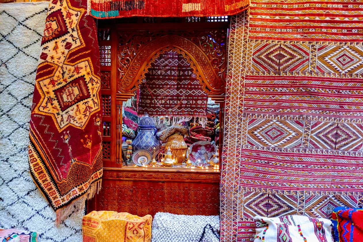 A vibrant Fes market stall displaying richly patterned Berber carpets and textiles, with carved wooden shelves filled with pottery, metalwork, and traditional crafts.