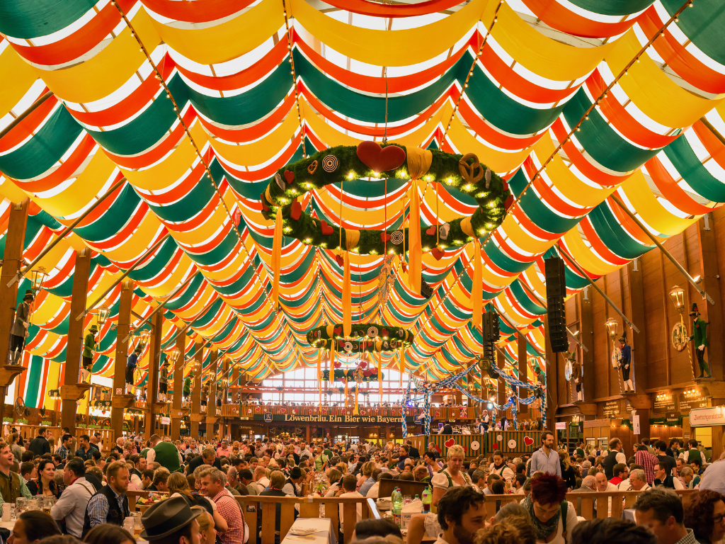 An Oktoberfest beer tent in Munich. Multi-colored awning and hundreds of people seated around tables enjoying beers and food.