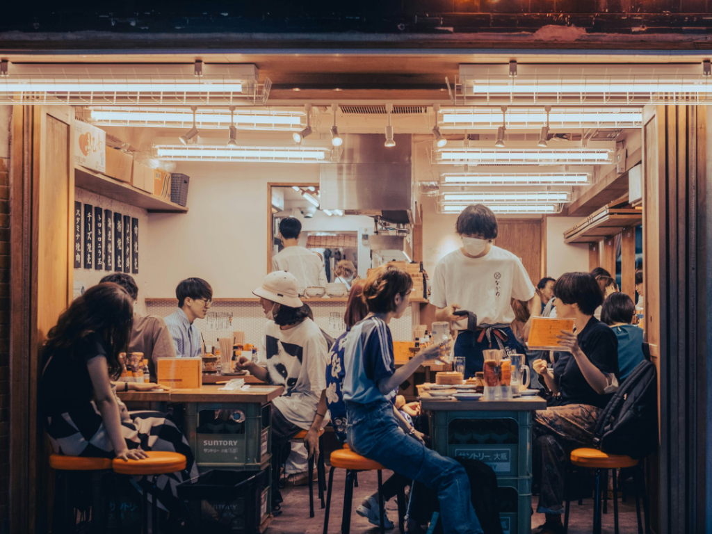 A shot of patrons sitting in a small izakaya during the evening. The table in the foreground is asking questions of the waiter.