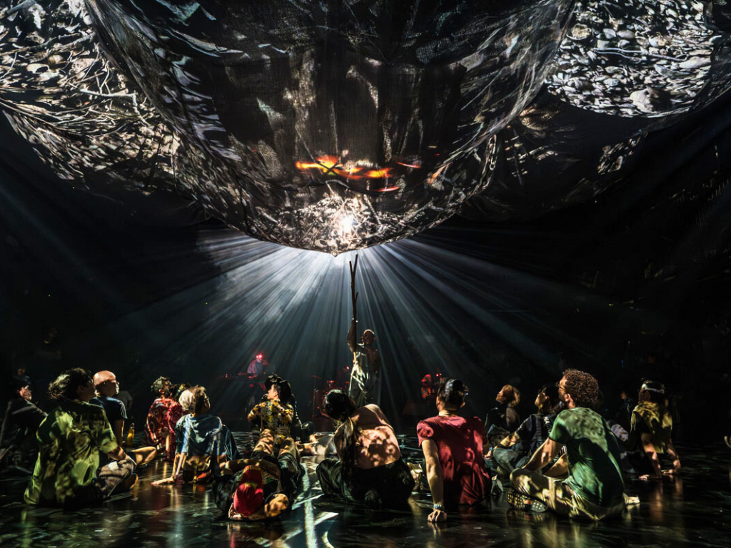 A group of performers reaching up toward a globe-like object in the ceiling during a performance at the Singapore International Festival of Arts.