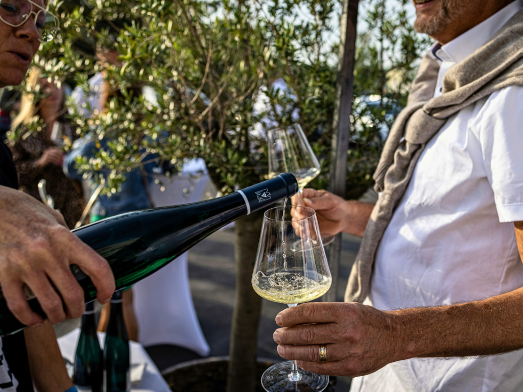 A waiter with black hair and glasses, slightly blurred and half off-frame, pours white wine into a glass held by a man in his fifties with a tan sweater draped over his shoulder. 