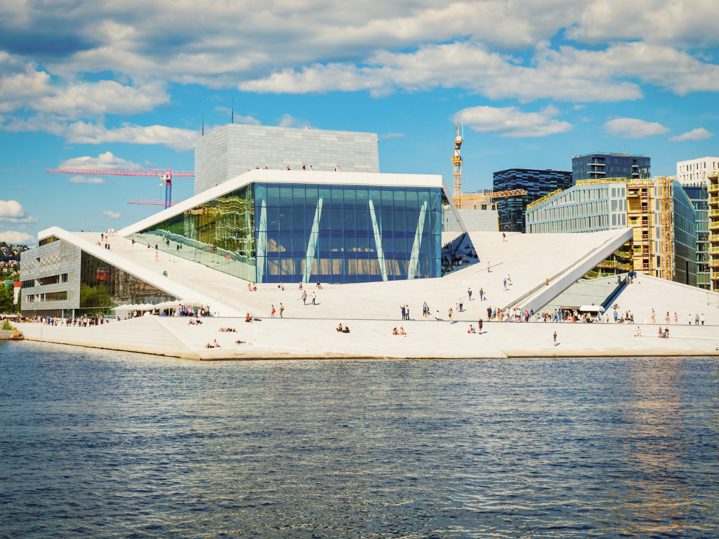 The Opera House in Oslo. An angular building set on the water, city buildings can be seen behind it. People dot the paved area in front of the Opera.