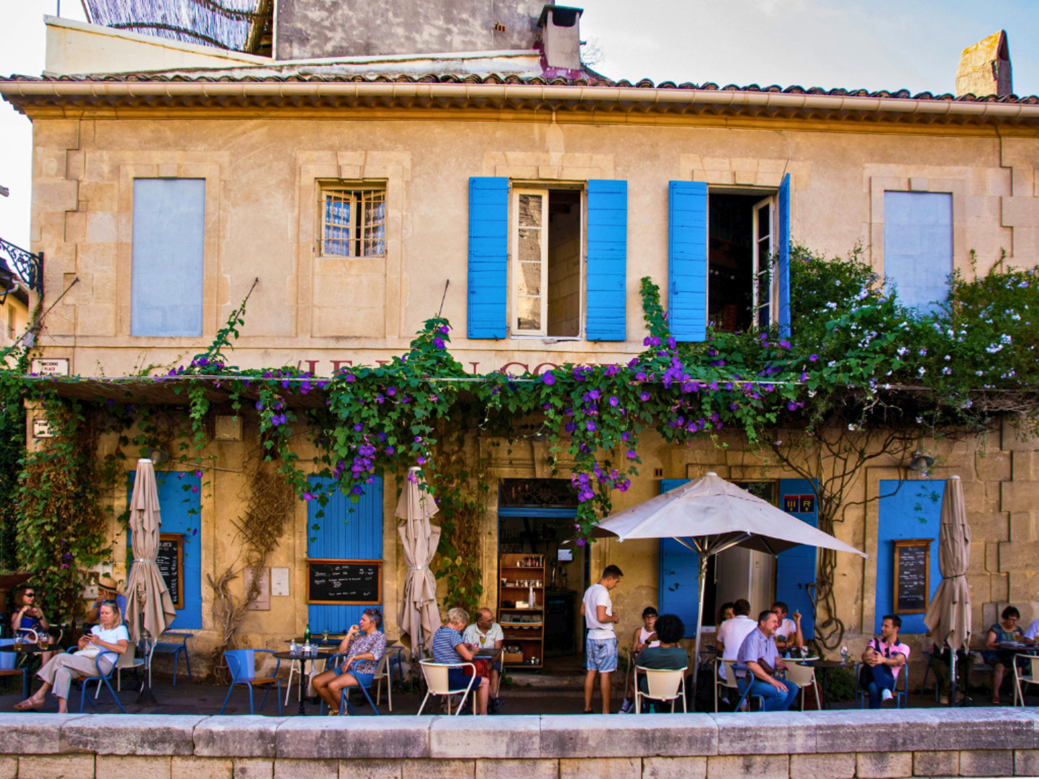 Small groups sit outside a cafe in Arles, a tan building with bright blue shutters. 