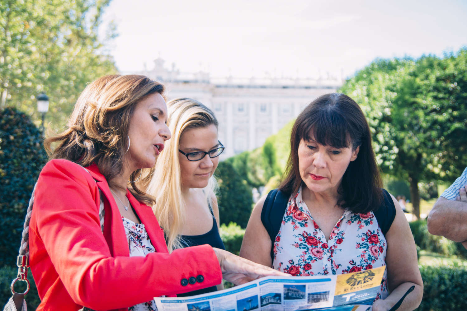 A guide in a bright red jacket points to a brochure, explaining something to two female travelers who are visiting Madrid. A nondescript building and green trees are blurred in the background. 