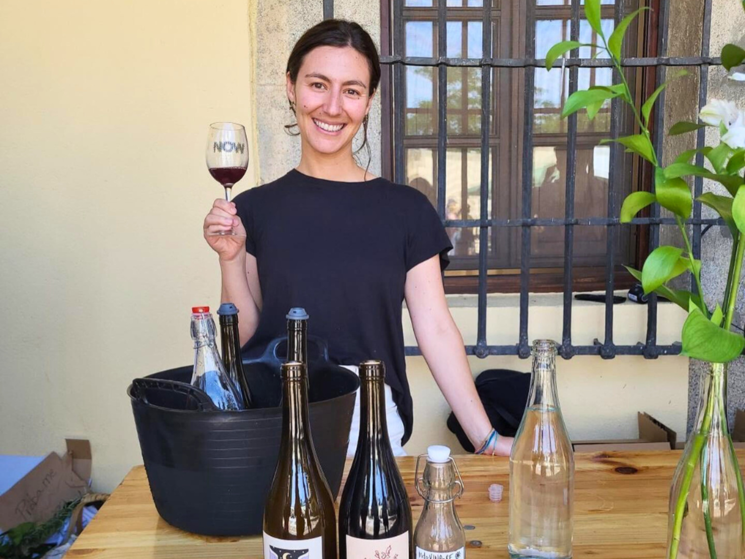 Nicole holds a glass of red wine, a wine bucket with three bottles sits on the table in front of her, and three bottles of her locally produced wine sit at the edge of the table.