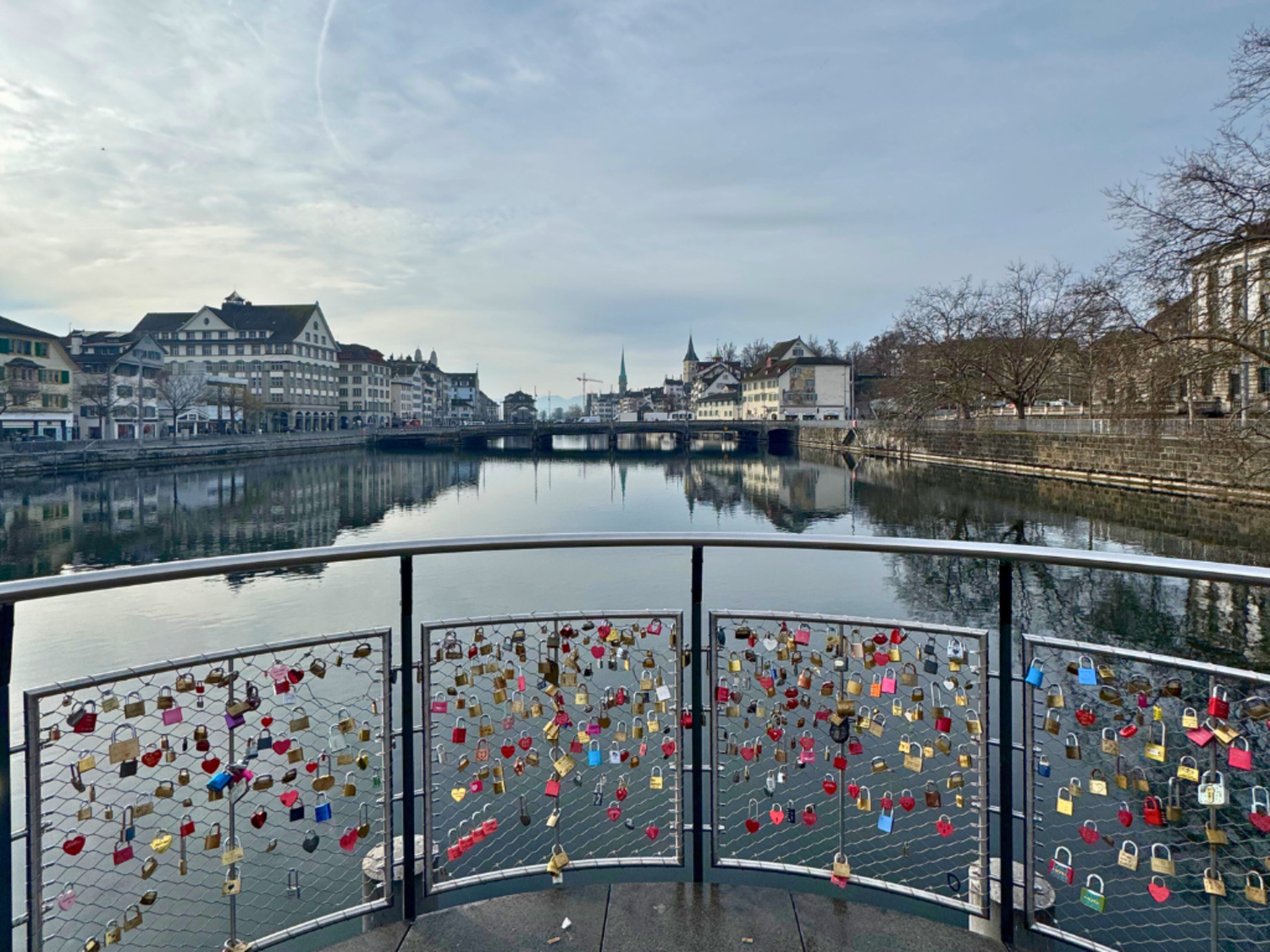 A view out over the water in Zurich. A fence with locks is in the foreground, the famous Mühlesteg bridge. 