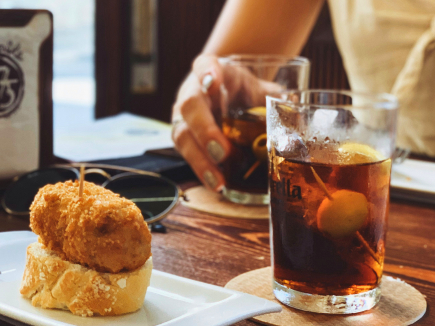 A croquette and glass of vermouth sit on a table, in the background a woman's hand holds another glass of vermouth.