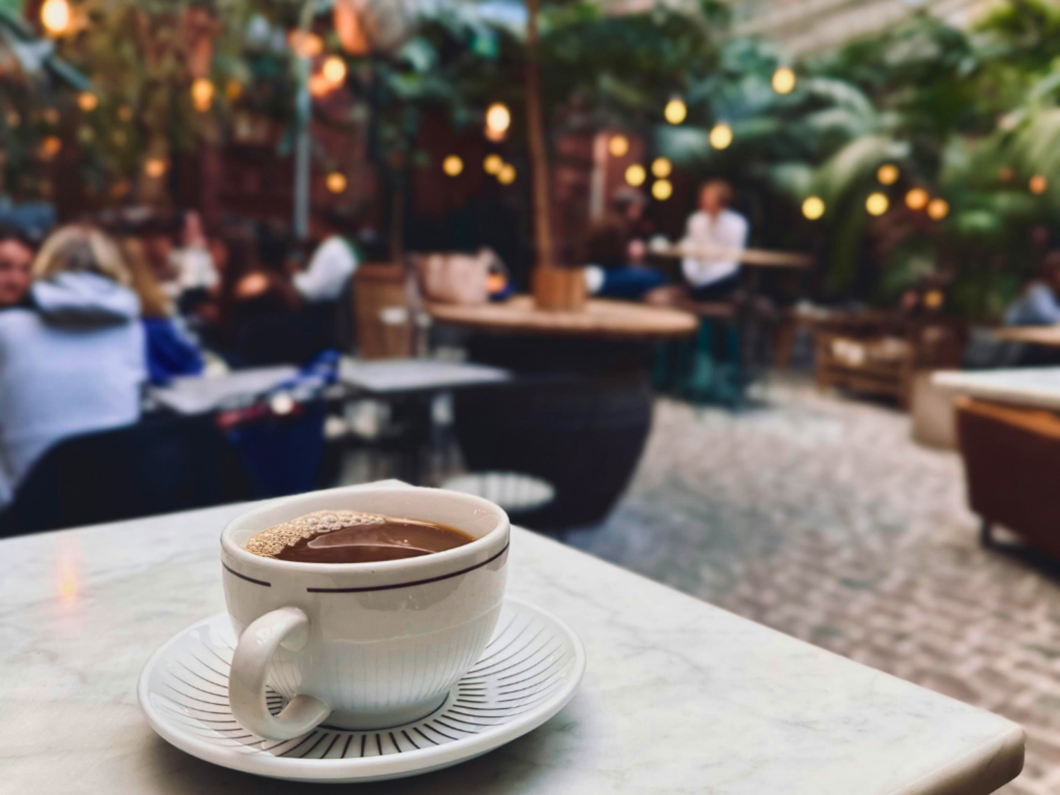 A coffee sits on a table, patrons in the background blurred out beneath bokeh lights.