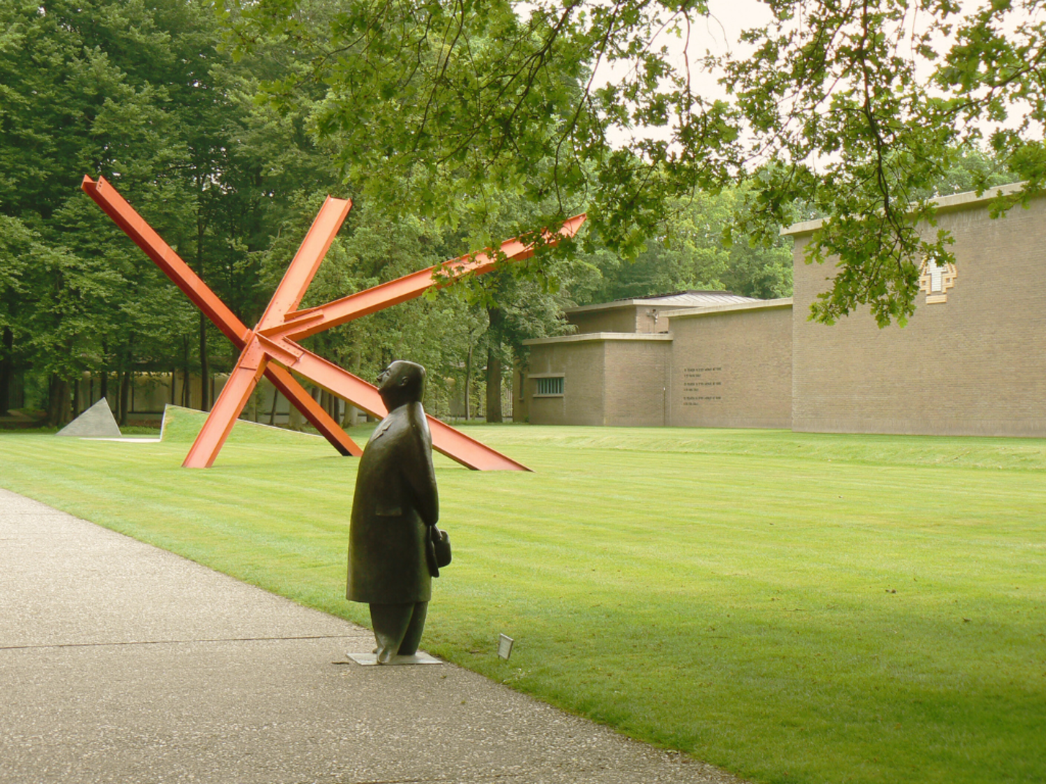 A sculpture stands in the foreground, in the background the red K-piece by Mark di Suvero. The Kroller Muller museum is behind both of them in the background. 