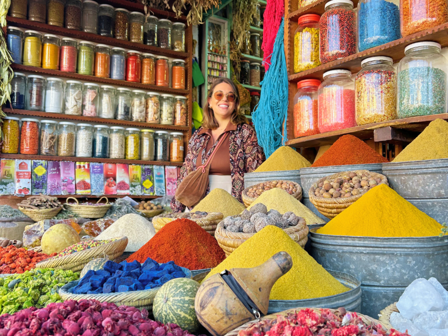 A woman stands in a spice stall in Marakech, colorful spices in baskets are in the foreground and jars of spices are on the wall behind the woman. 