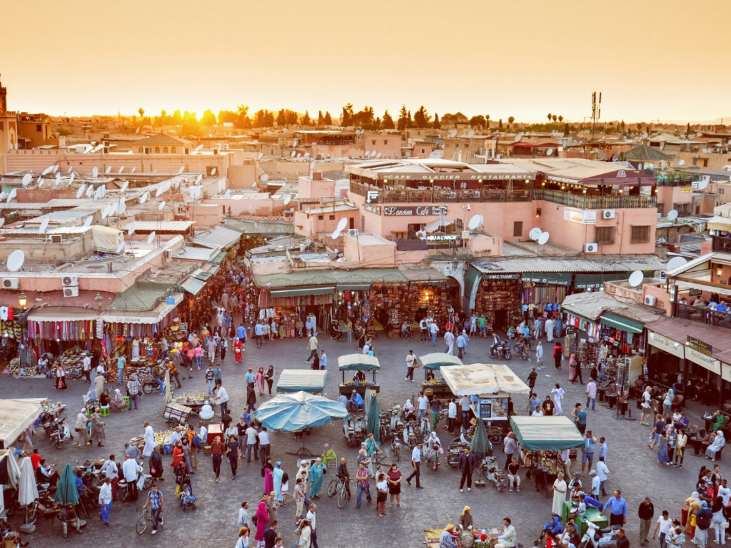 A low aeiral view of the sun setting with buildings are in the background. In the foreground is a public square with market stalls and many people shopping.