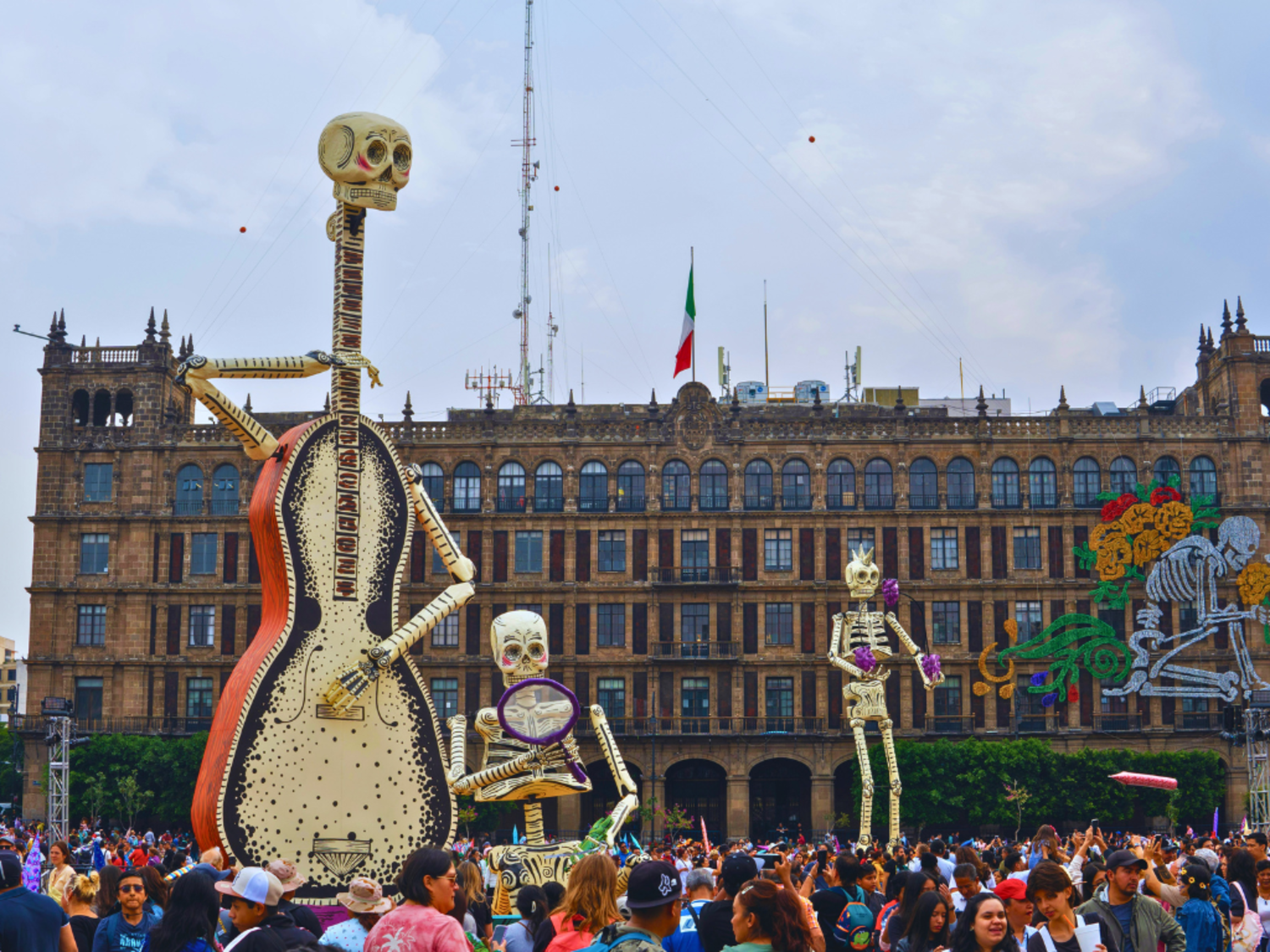 A crowd gathers for Dia de los Muertos celebration, three giant skeletons tower over the people.