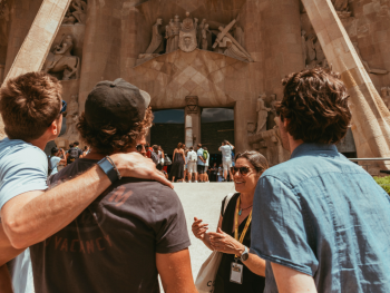 Three guests with backs to the camera look up at the Sagrada Familia, and an expert is facing them and the camera, explaining the facade to the travelers.