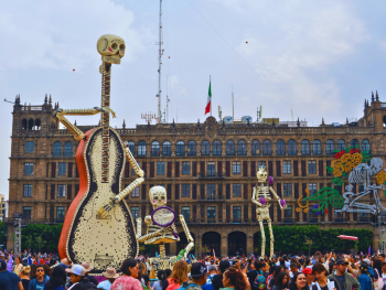 A crowd gathers for Dia de los Muertos celebration, three giant skeletons tower over the people.